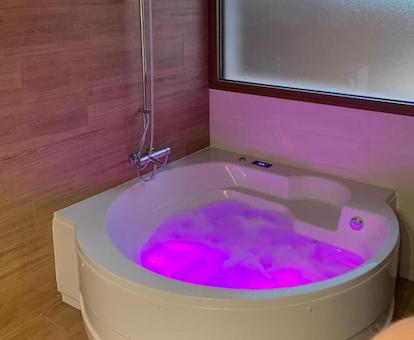 Jacuzzi circular con cromoterapia y en color rosa en el cuarto de baño junto a la pared y debajo de una ventana biselada.