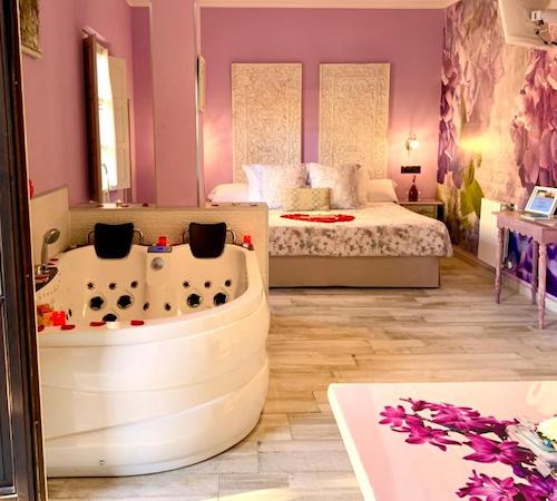 Jacuzzi para dos personas con chorros de agua para masaje en la espalda en una habitación con la cama al fondo y pintada de color de rosa en la suite con bañera de hidromasaje del hotel Suites con Jacuzzi Santillana en Santillana del Mar